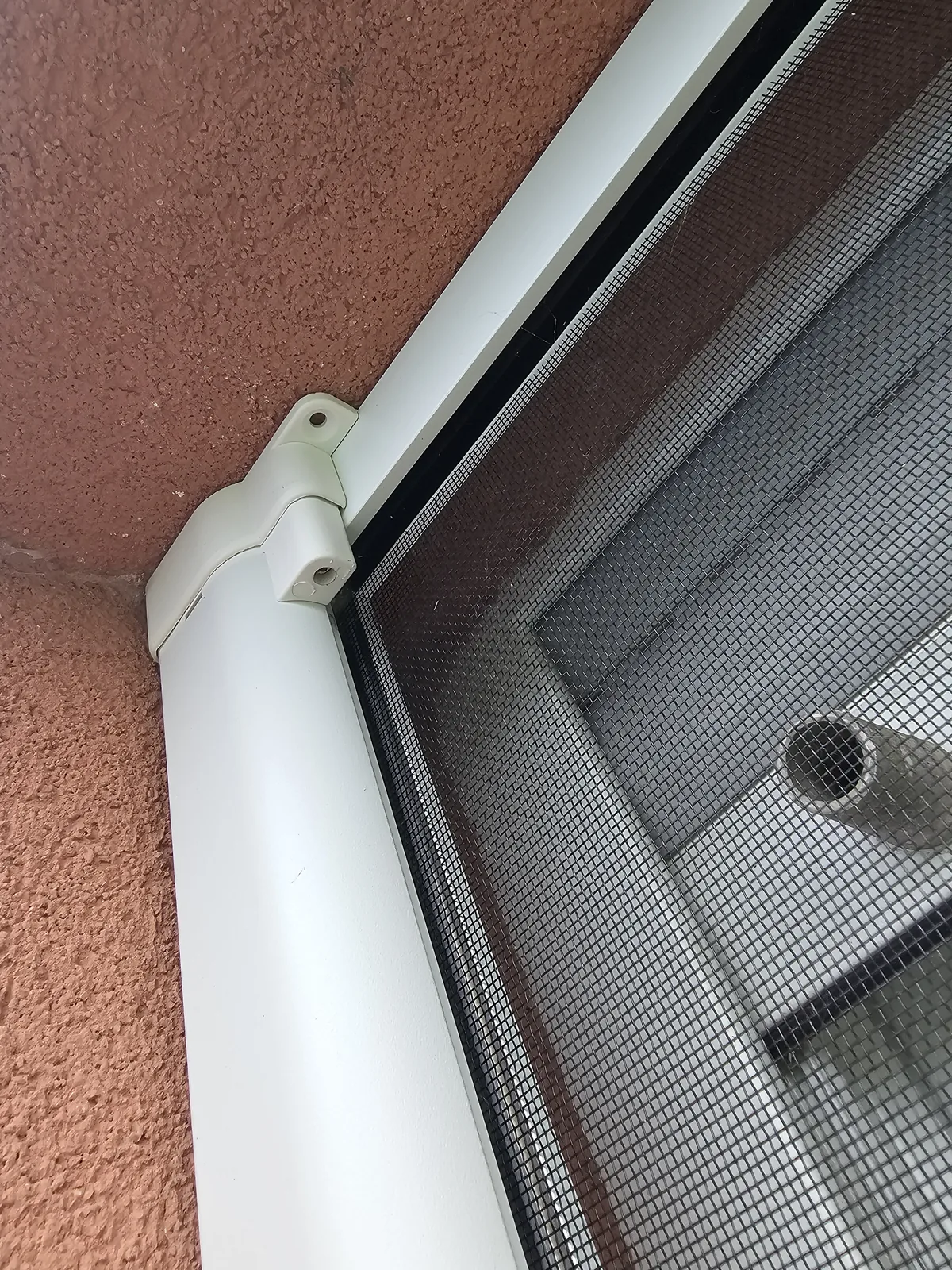 Support blanc vissé en façade qui tient le rail haut de la moustiquaire coulissante, toile visible derrière.