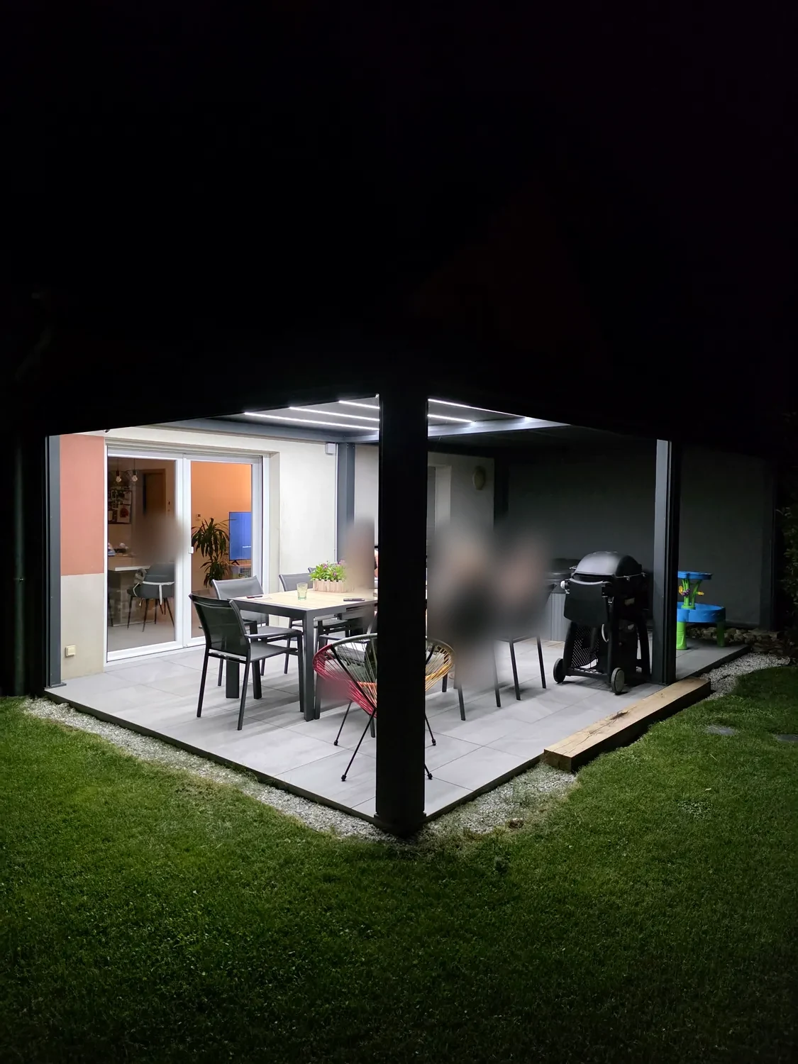 Pergola bioclimatique anthracite de nuit, lames fermées et LED allumées au-dessus d’une table dressée sur une terrasse dallée.