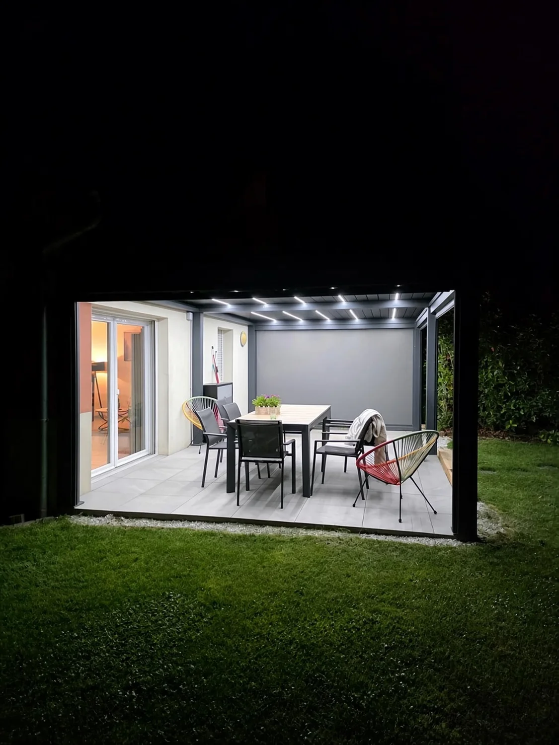 Pergola bioclimatique anthracite de nuit, LED allumées dans les lames, store baissé au fond, table et chaises sur une terrasse dallée.