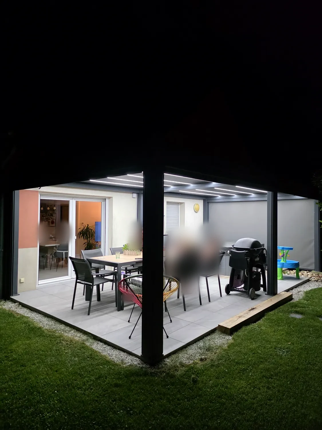 Invités attablés sous une pergola bioclimatique éclairée par les LED intégrées aux lames, terrasse dallée, baie vitrée ouverte sur la maison.